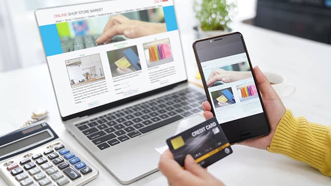 Casi todas las actividades en línea pueden contribuir a tu huella digital. La imagen muestra a alguien sosteniendo su teléfono y su tarjeta de crédito frente a la pantalla de una computadora con una ventana de compras en línea abierta.